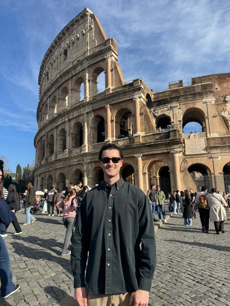 Man in dark shirt and sunglasses standing in front of the ancient Roman Colosseum in Rome, Italy on a sunny day with tourists