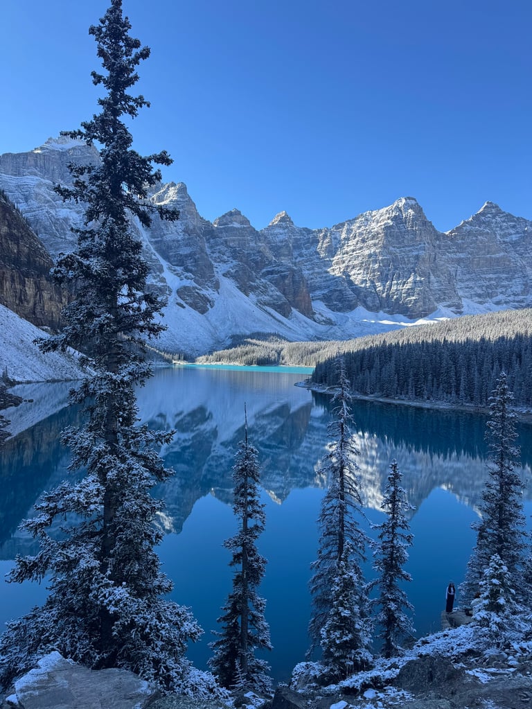 Snow-covered evergreen trees frame a pristine alpine lake with turquoise water and snow-capped mountain peaks reflected in the background