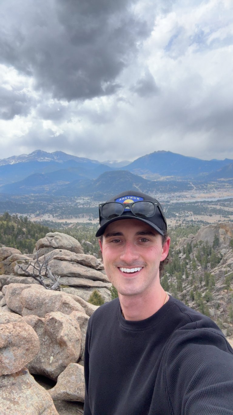Man smiling at camera on rocky mountain overlook with distant mountains and valley landscape under cloudy sky
