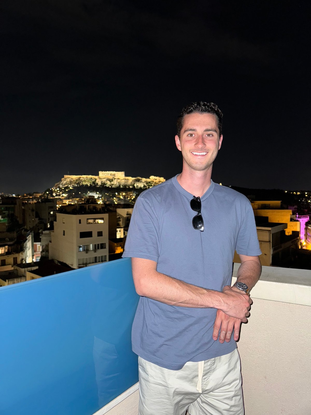 Man in blue shirt smiling at rooftop bar with illuminated Acropolis visible at night in Athens background