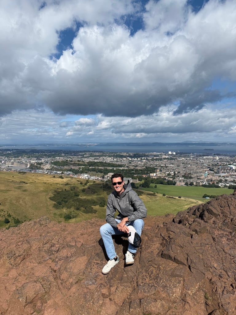 Man in sunglasses crouching on rocky hillside overlooking a sprawling city valley with green fields and cloudy sky