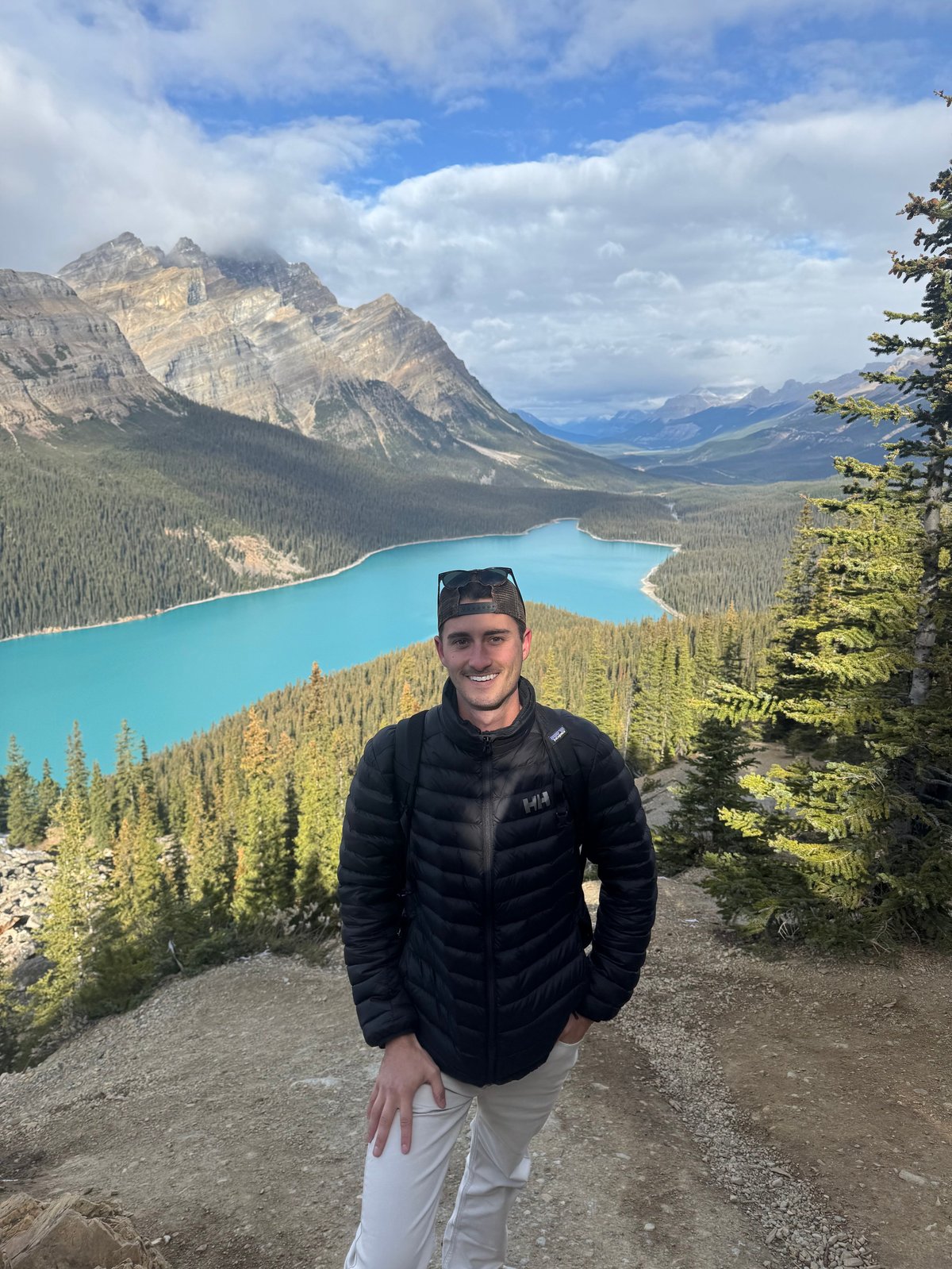 Man smiling at camera with turquoise lake, mountains, and forested valley in background