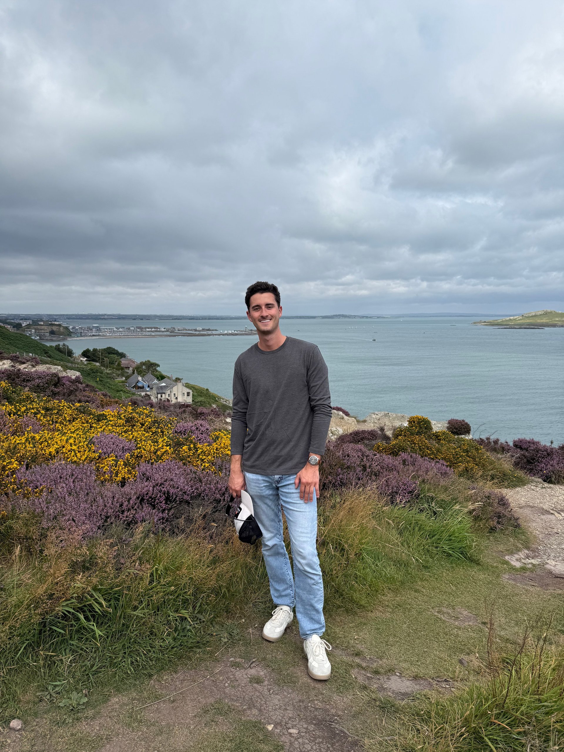 Man standing on coastal cliff with purple heather flowers, overlooking blue sea and cloudy sky