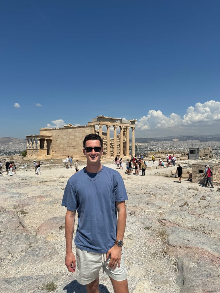 Man in blue shirt and sunglasses smiling at ancient Greek temple ruins with tourists and cityscape in background