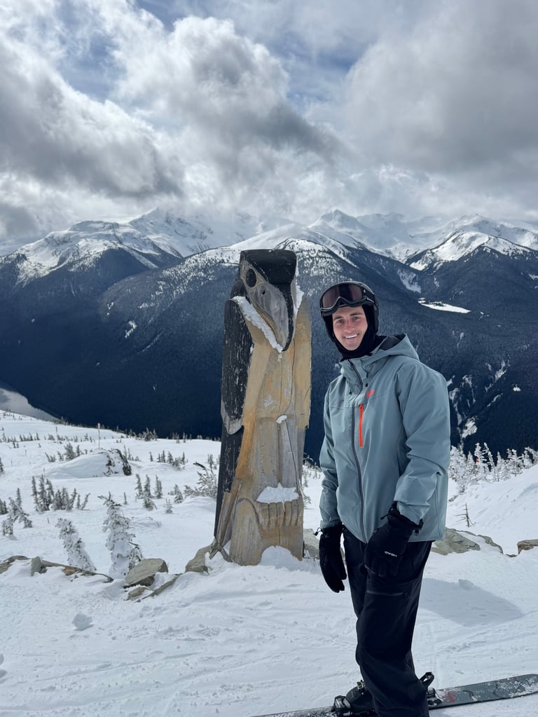 Person in winter gear posing next to carved wooden figure with snowy mountains and forest in background