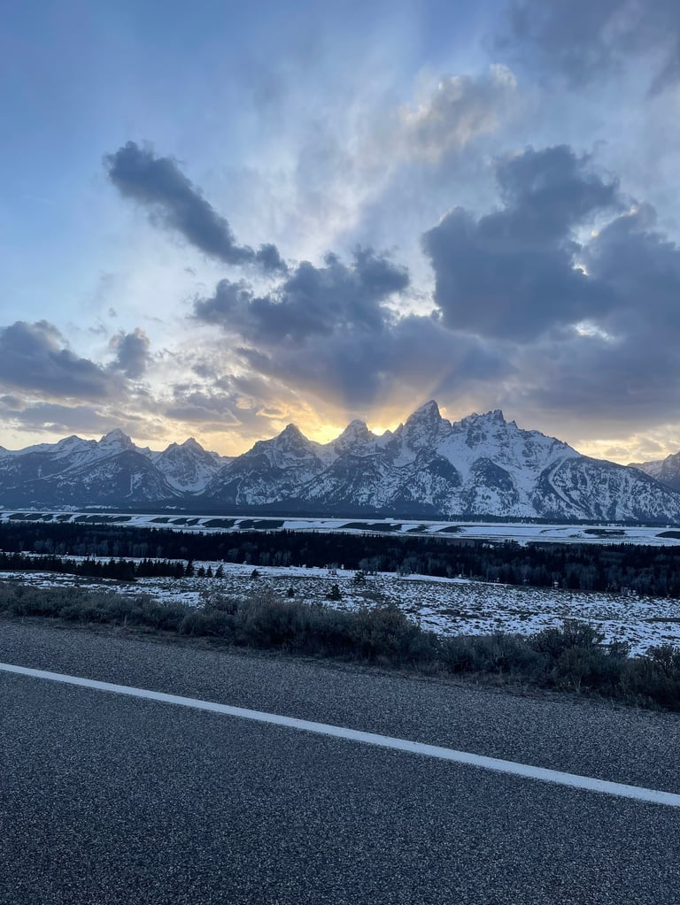 Snow-covered mountain peaks at sunset with dramatic storm clouds overhead and a paved road in the foreground