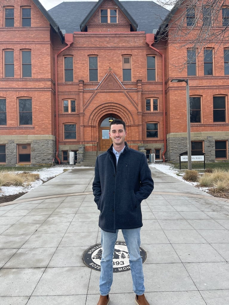 A man in a dark jacket and jeans stands on a stone plaza in front of a large red brick historic building with arched entrance