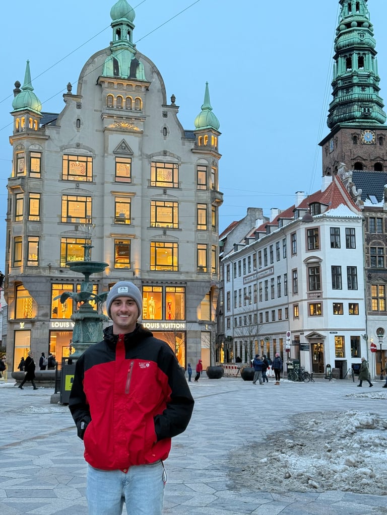 Person in red jacket standing in historic European city square at dusk with illuminated buildings